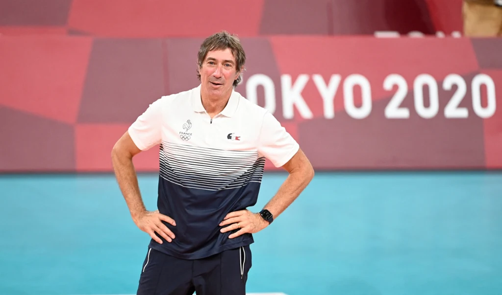 Laurent Tillie Trener w sportowej koszulce i spodenkach stoi na tle symboli igrzysk olimpijskich Tokio 2020, uśmiechnięty, z rękami opartymi na biodrach, w hali sportowej.