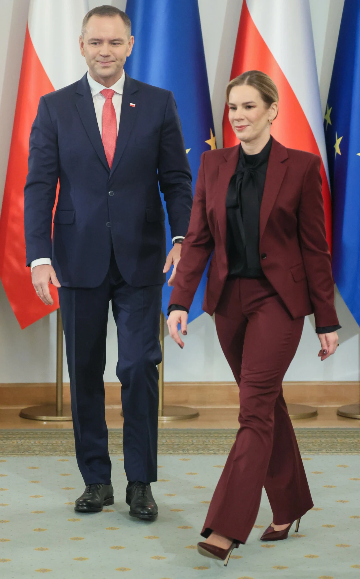 Dobrze skrojony damski garnitur jest obecnie hitem od kilku miesięcy  Dwie osoby w eleganckich garniturach kroczące po jasnej podłodze w pomieszczeniu z flagami Polski i Unii Europejskiej w tle.