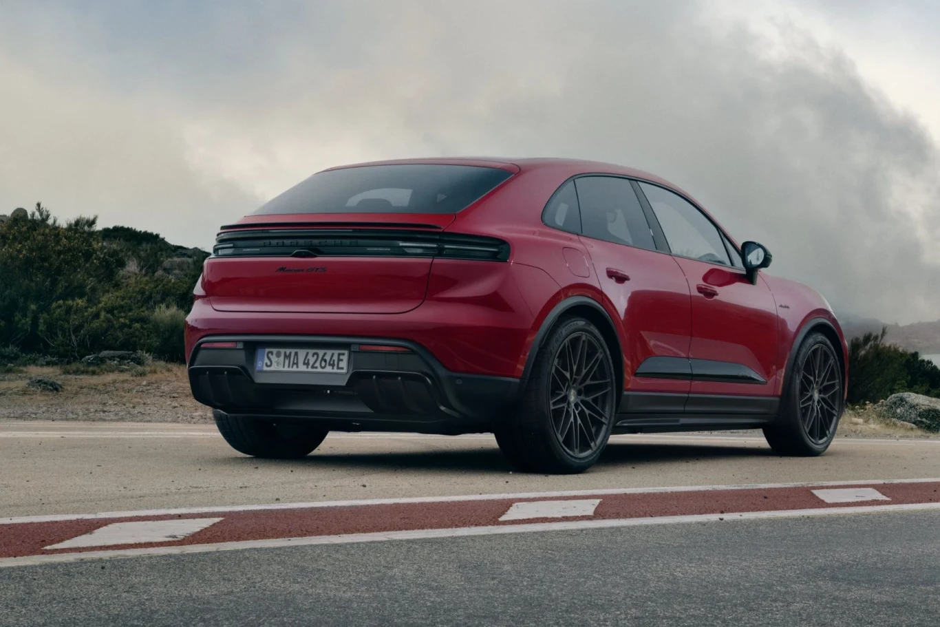 Standardowo Macan GTS wyposażony jest w 21-calowe felgi. Opcjonalnie można zdecydować się na felgi 22-calowe. Czerwony sportowy SUV zaparkowany przy drodze, widok z tyłu, dach opadający w stylu coupé, masywne nadkola i eleganckie felgi, w tle niska roślinność i zachmurzone niebo.