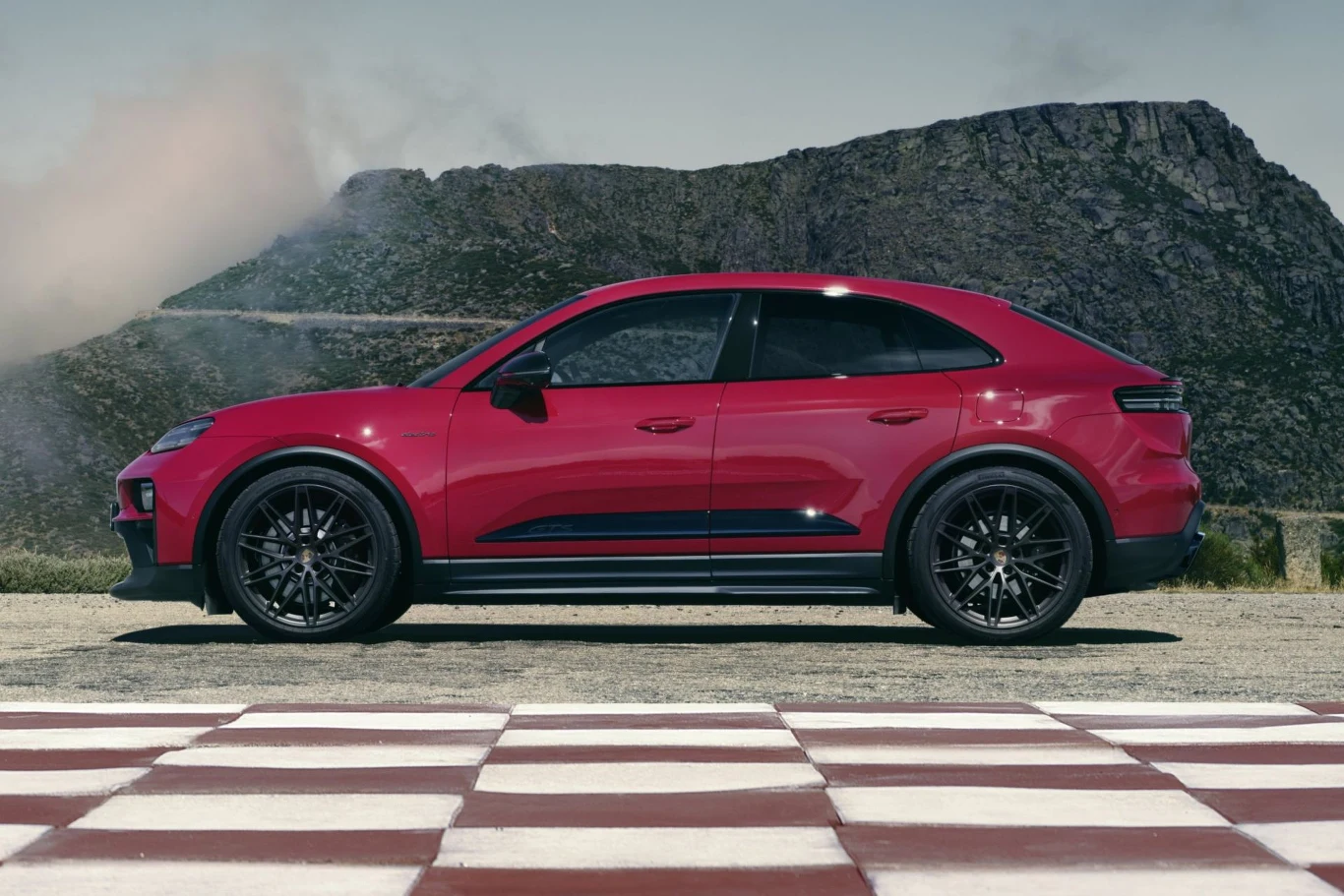 Rozkład masy w Porsche Macanie GTS wynosi 48:52. Czerwony samochód sportowy zaparkowany na tle górskiego krajobrazu, widoczny z profilu, asfaltowa nawierzchnia z białymi i czerwonymi pasami na pierwszym planie, w tle szczyt górski i czyste niebo.