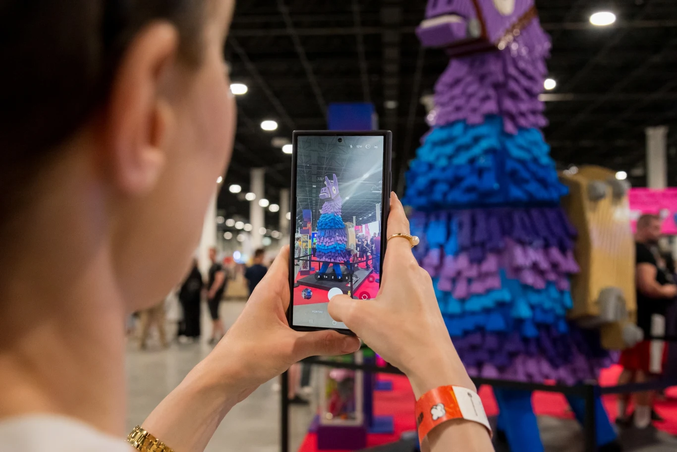 Lama Zaopatrzeniową Lego Fortnite Osoba fotografuje kolorową figurę lamy ozdobioną fioletową i niebieską bibułą na tle przestronnej hali wystawienniczej.