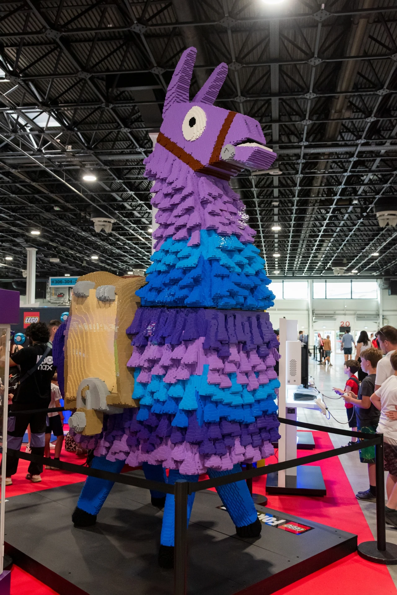 Duża, kolorowa figura lamy zbudowana z klocków LEGO, otoczona barierkami i odwiedzana przez ludzi w przestronnej hali wystawowej.