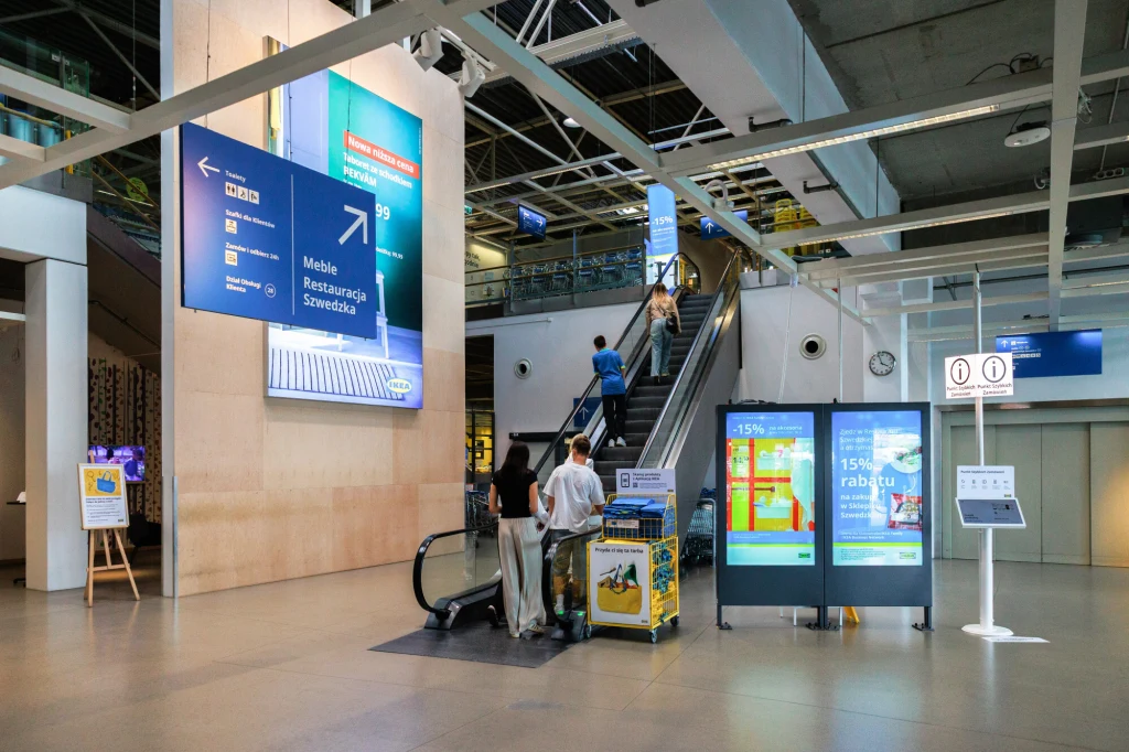 Ikea Wnętrze dużego sklepu meblowego z widocznymi schodami ruchomymi, dużymi kierunkowskazami informacyjnymi, osobami z wózkami sklepowymi i elektronicznymi tablicami reklamowymi.