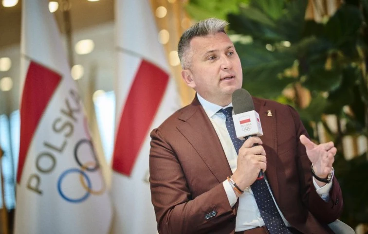 Radosław Piesiewicz, źr. PKOl Mężczyzna w brązowym garniturze trzymający mikrofon z logo TVP Sport, udzielający wywiadu lub wypowiedzi na tle flag z napisem 'Polska' i symbolem olimpijskim, w otoczeniu roślin.