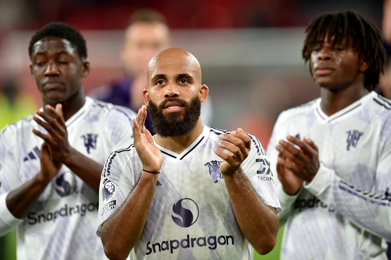 Manchester United Trzech piłkarzy w białych koszulkach sportowych z logo Snapdragona bije brawo po meczu na stadionie, centralnie znajduje się łysy zawodnik z brodą, a po bokach stoją jego koledzy z drużyny.