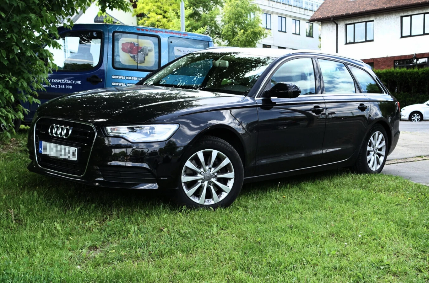 Czarny samochód osobowy marki Audi zaparkowany na zielonej trawie w pobliżu chodnika, w tle widoczny niebieski pojazd użytkowy oraz fragment budynku mieszkalnego.