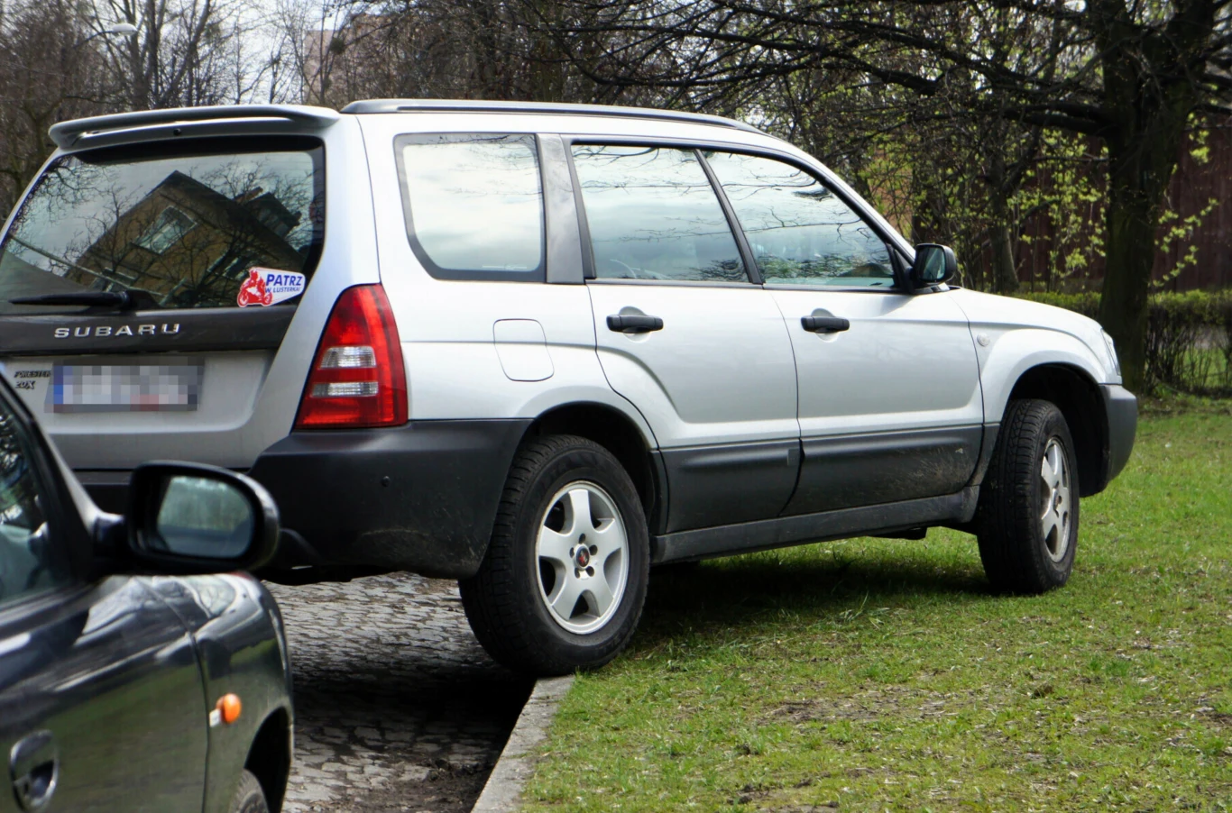 Ile wynosi mandat za parkowanie na trawniku? Srebrny samochód Subaru częściowo zaparkowany na chodniku oraz trawie, tyłem do krawężnika, w otoczeniu drzew i drugiego pojazdu.