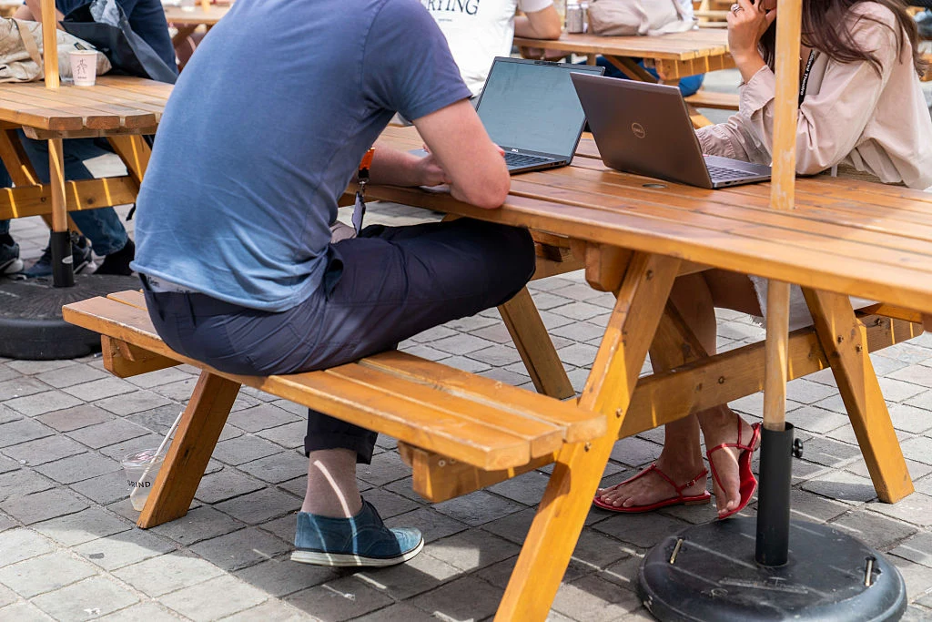 Dwie osoby siedzą przy drewnianym stole piknikowym na zewnątrz, skupione na pracy przy laptopach; widoczne nogi i ubrania, fragmenty kubków z napojami oraz brukowany plac.
