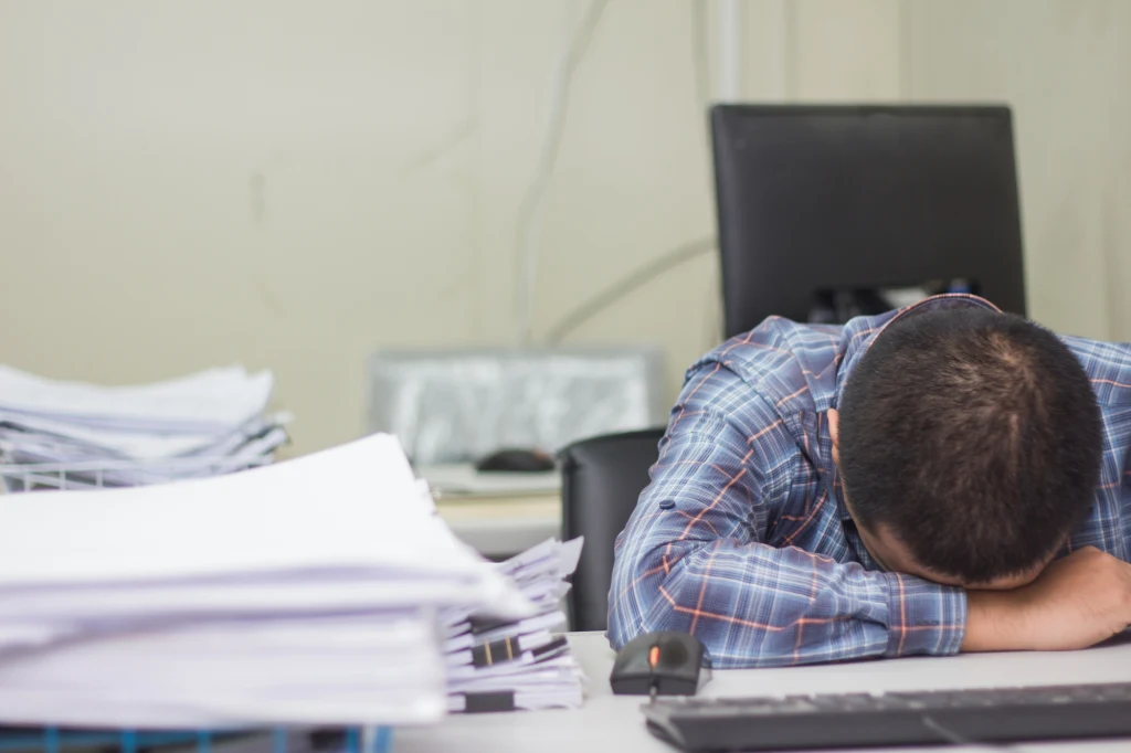 Mężczyzna opiera głowę na biurku w pobliżu stosu dokumentów oraz klawiatury, wyrażając zmęczenie lub frustrację związane z pracą biurową.