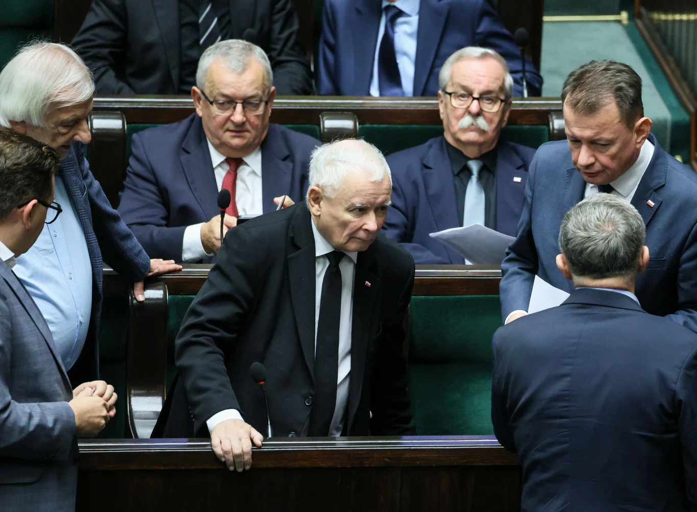 Grupa polityków w garniturach podczas dyskusji w sali sejmowej, w centrum uwagi stoi mężczyzna z siwymi włosami w czarnym garniturze, wokół niego inni parlamentarzyści, atmosfera skupienia