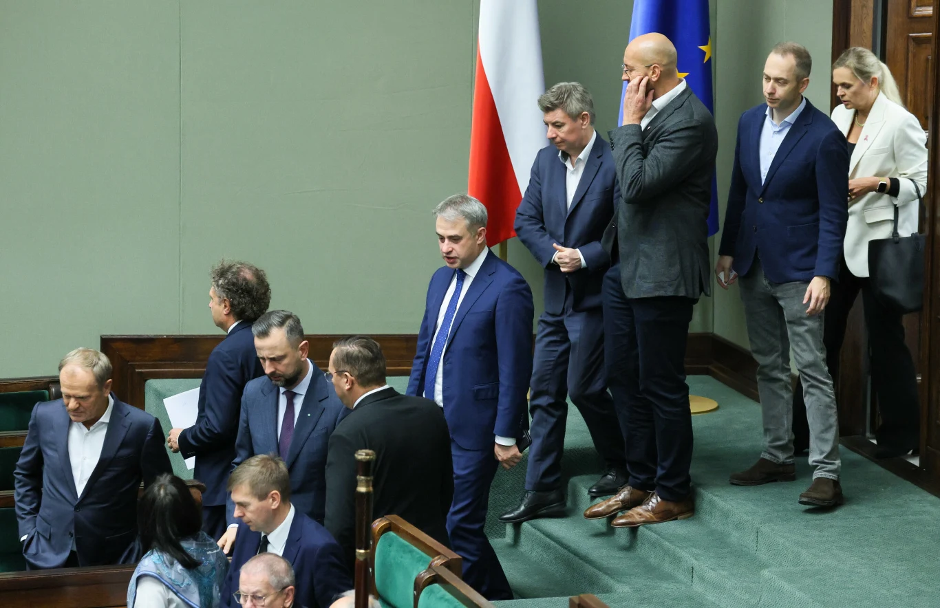 Grupa polityków opuszcza salę obrad sejmowych, wewnątrz widoczne są flagi Polski oraz Unii Europejskiej, wszyscy ubrani są elegancko w formalne stroje, atmosfera poważna i skupiona.