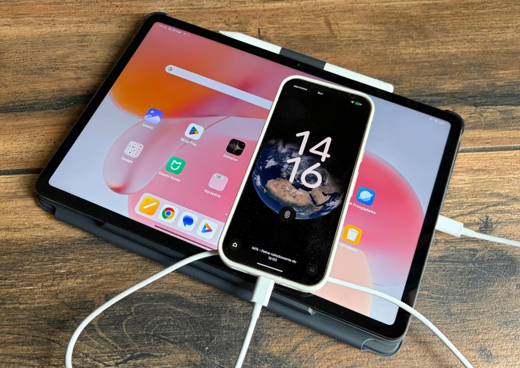 Redmi Pad 2 Pro pozwala na szybkie ładowanie baterii innych urządzeń. Tablet z dużym ekranem i uruchomionymi aplikacjami leży na drewnianym blacie, na jego środku znajduje się podłączony białym kablem smartfon wyświetlający ekran blokady z godziną oraz zdjęciem Ziemi, oba urządzenia ładują się jednocześnie.