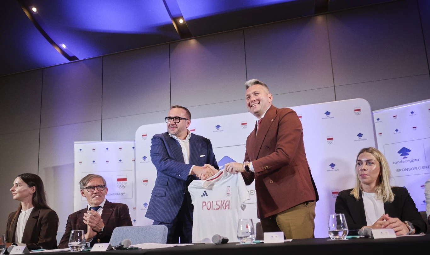 Dwóch mężczyzn w eleganckich garniturach ściska sobie dłonie, trzymając koszulkę z napisem Polska, otoczeni przez trzy inne osoby siedzące przy stole konferencyjnym, w tle bannery z logotypami oraz symbolem książki.