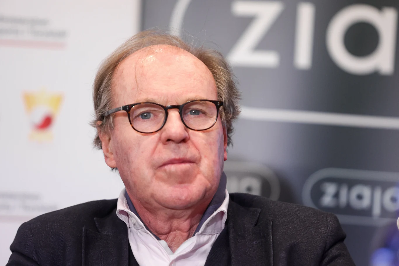 Starszy mężczyzna w okularach, ubrany w marynarkę i koszulę, siedzi na tle rozmytego napisu oraz logotypów, patrząc prosto przed siebie. Wyraz twarzy poważny, neutralne światło, tło konferencyjne.