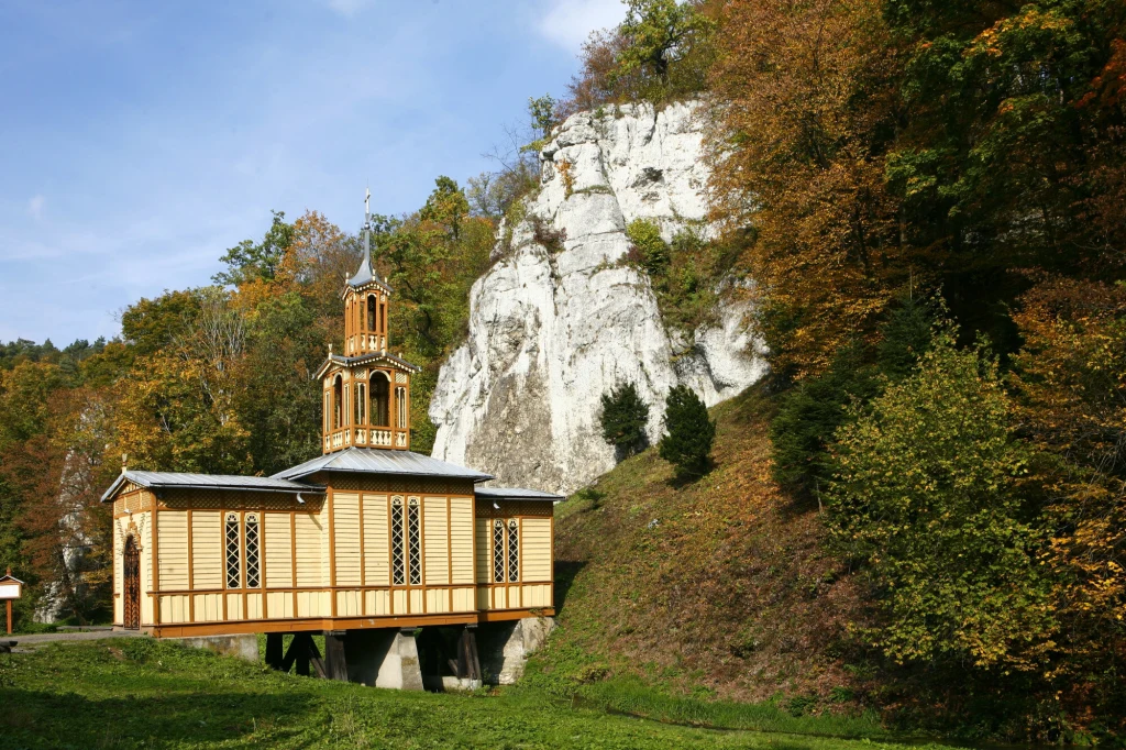 Drewniany kościółek z wieżyczką na tle białych wapiennych skał, otoczony jesiennym lasem, błękitne niebo i soczysta zieleń trawy podkreślają malowniczość krajobrazu.