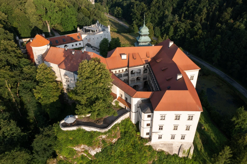 Zabytkowy zamek z jasnymi murami i czerwonymi dachami otoczony gęstymi drzewami, widoczny z lotu ptaka, z tarasem na skarpie oraz wieżyczkami i charakterystyczną kopułą w tle.
