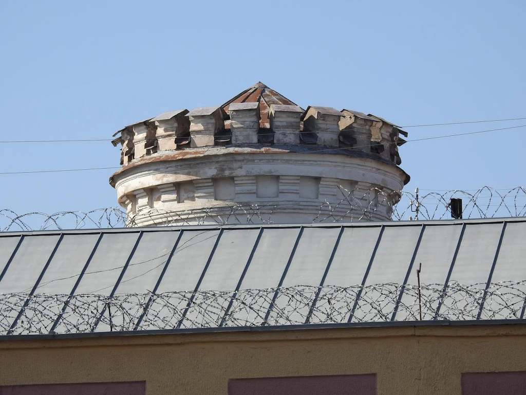 Stara wieża strażnicza z cegły z daszkiem, otoczona ogrodzeniem z drutu kolczastego, na tle nieba bez chmur.