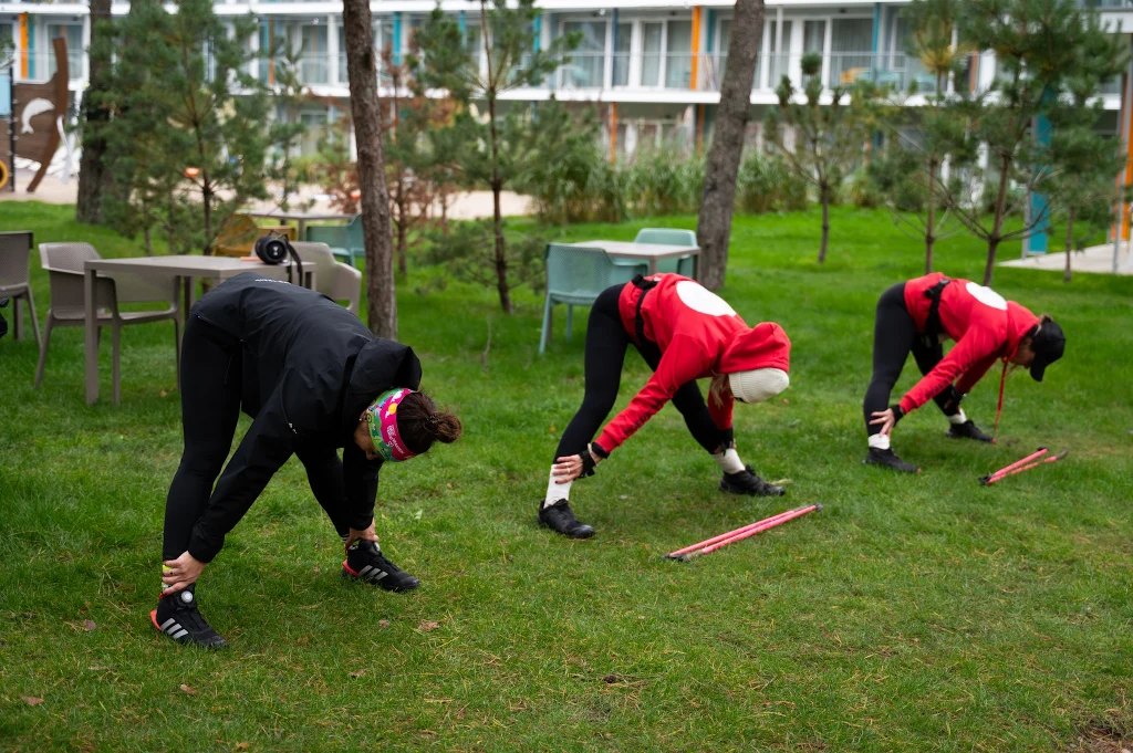 Trzy osoby w odzieży sportowej wykonujące rozciąganie na trawniku, każda z nich pochyla się do przodu i trzyma za kostki, w pobliżu leżą kijki do nordic walking, w tle widoczne są drzewa, zielona trawa i meble ogrodowe.