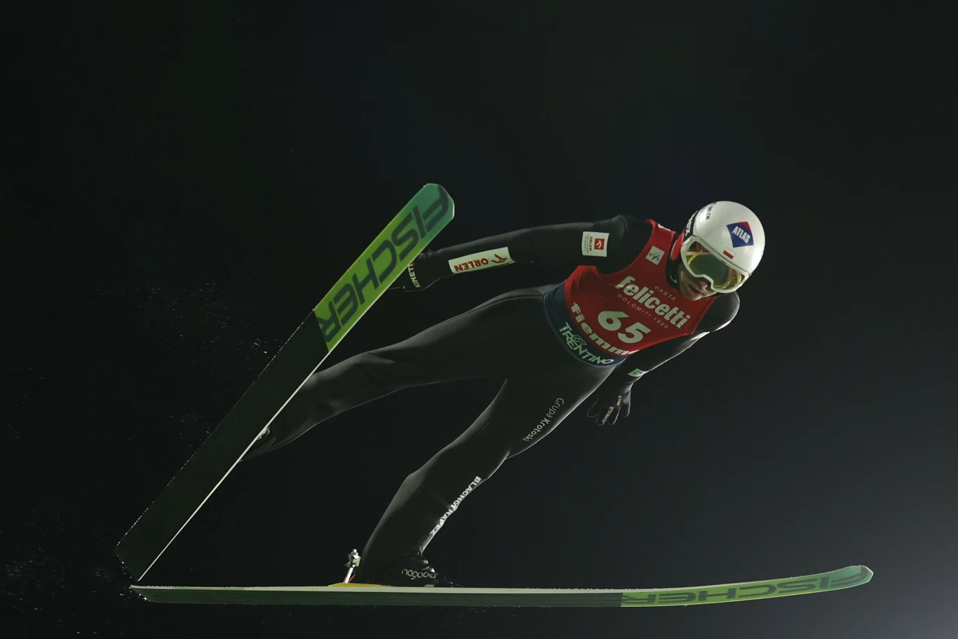Kamil Stoch na skoczni zimowych igrzysk olimpijskich 2026 w Predazzo Skoczek narciarski w powietrzu podczas lotu, ubrany w czarny kombinezon, czerwony numer startowy i biały kask, trzyma narty ułożone równolegle w nocy na tle ciemnego nieba.