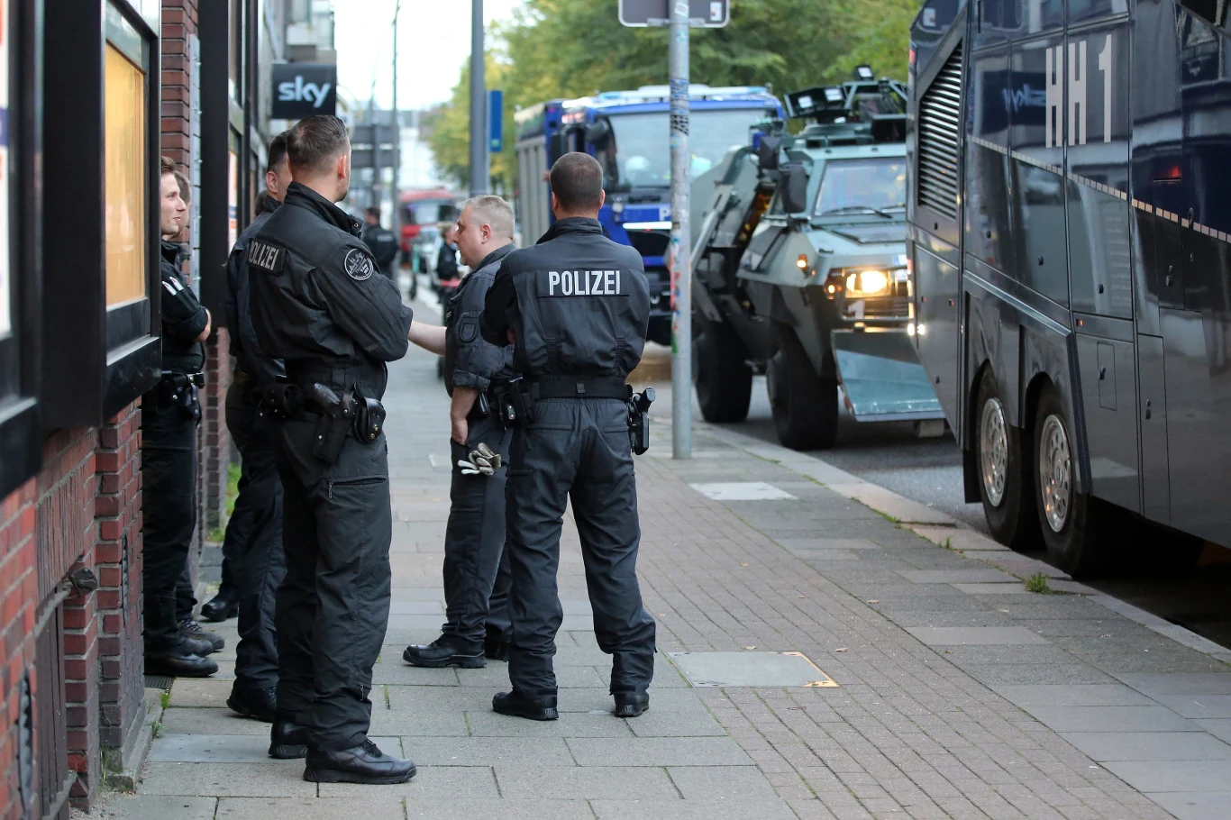 Hamburska policja przed mieszkaniem Olafa Scholza. Miasto płaci za to dwa miliony rocznie Grupa uzbrojonych funkcjonariuszy niemieckiej policji stojąca na chodniku obok policyjnego pojazdu opancerzonego i autobusu, w tle widać ulicę z zaparkowanymi pojazdami służbowymi.