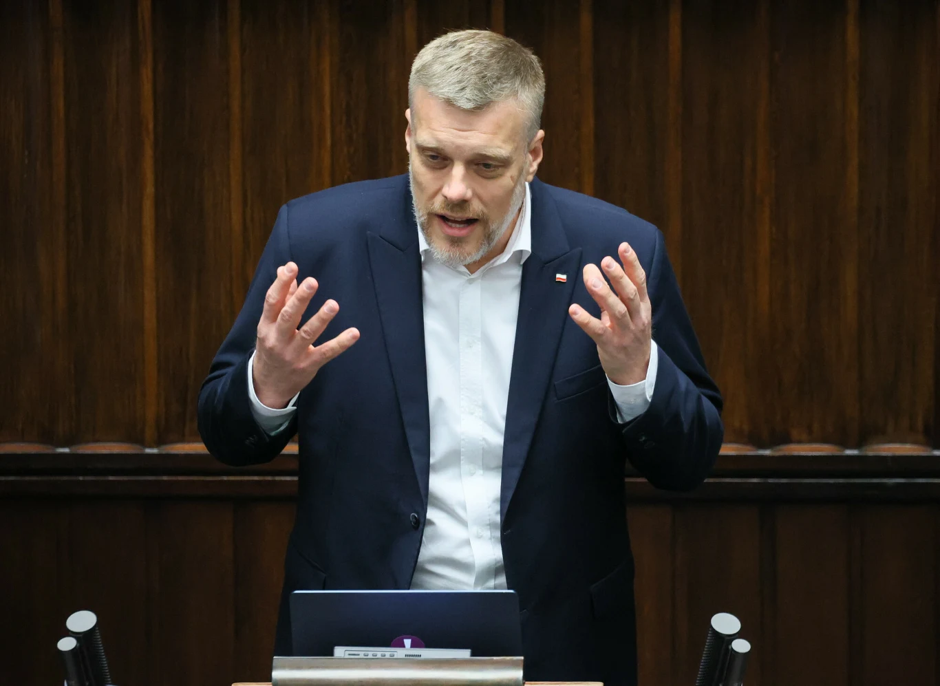Adrian Zandberg chce podatku od pustostanów deweloperskich Mężczyzna w marynarce i jasnej koszuli gestykulujący w trakcie przemówienia na mównicy, w tle drewniane panele charakterystyczne dla sali sejmowej.
