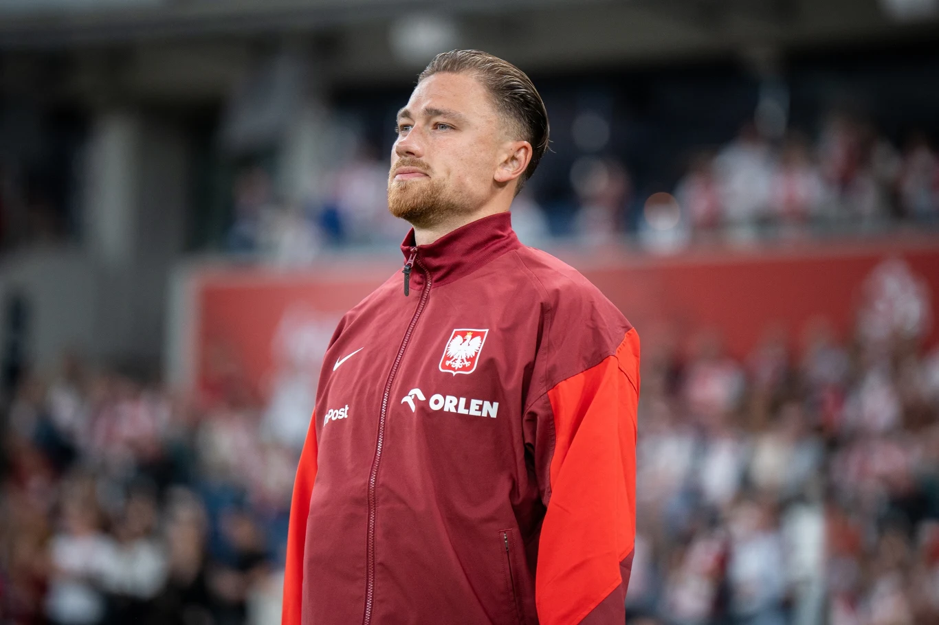 Mężczyzna w czerwonej kurtce reprezentacji Polski stoi na tle rozmytego stadionu, wśród widowni, z poważnym wyrazem twarzy, skupiony i pewny siebie, widoczne logo Orlen i godło Polski na kurtce.