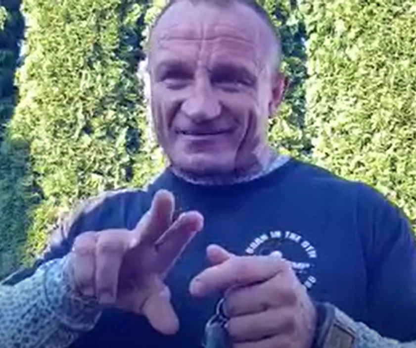 Mężczyzna z umięśnioną sylwetką uśmiecha się, trzymając w dłoniach przedmiot przypominający klucze, w tle zielony żywopłot.