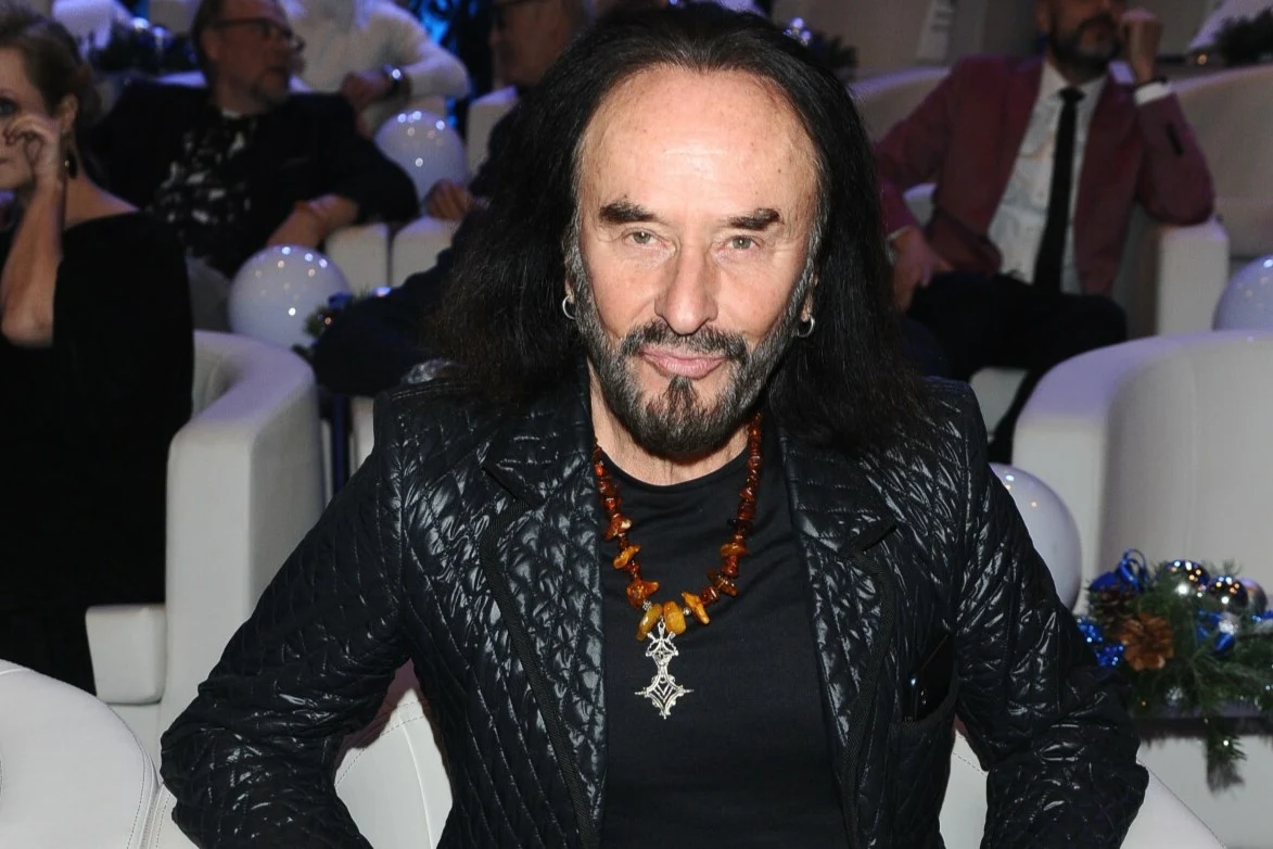 Stan Borys posługuje się pseudonimem artystycznym Mężczyzna z długimi czarnymi włosami i brodą siedzi w jasnym fotelu, ubrany w czarną kurtkę z pikowaną fakturą i bursztynowy naszyjnik z krzyżem, w tle widoczni inni ludzie oraz balony dekoracyjne.