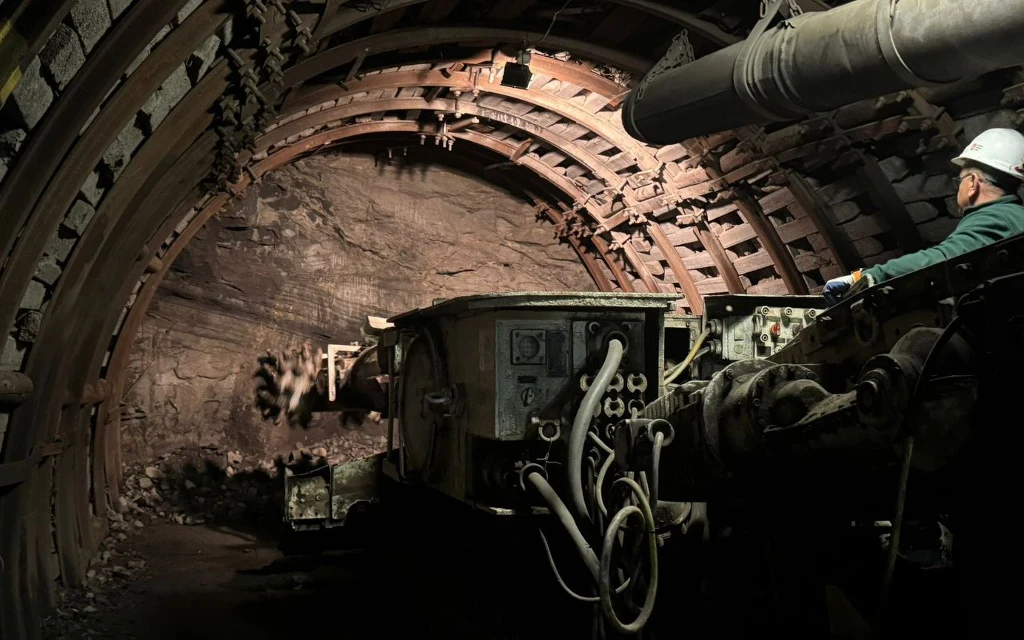 Górnik w kasku obsługuje ciężki sprzęt górniczy w oświetlonym tunelu wydrążonym w skale, sklepienie zabezpieczone jest metalowymi konstrukcjami, a ściany wykazują ślady eksploatacji surowców.