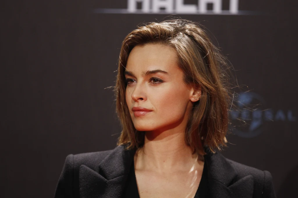 Młoda kobieta o brązowych włosach i jasnej karnacji w eleganckiej, czarnej marynarce na ciemnym tle z rozmytym napisem w górnej części kadru, wyraz twarzy poważny i skupiony.