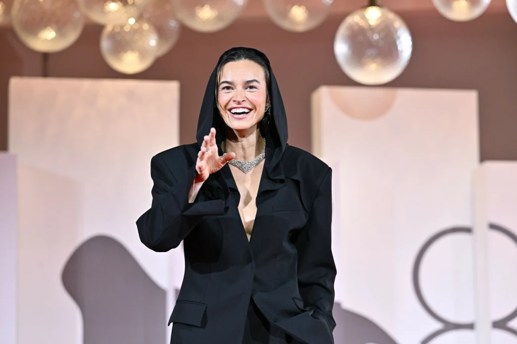 Uśmiechnięta kobieta w czarnym, eleganckim stroju z kapturem, nosząca okazały naszyjnik, gestykuluje dłonią w stronę obiektywu; za nią znajdują się dekoracyjne, jasne elementy scenografii oraz wiszące kule.