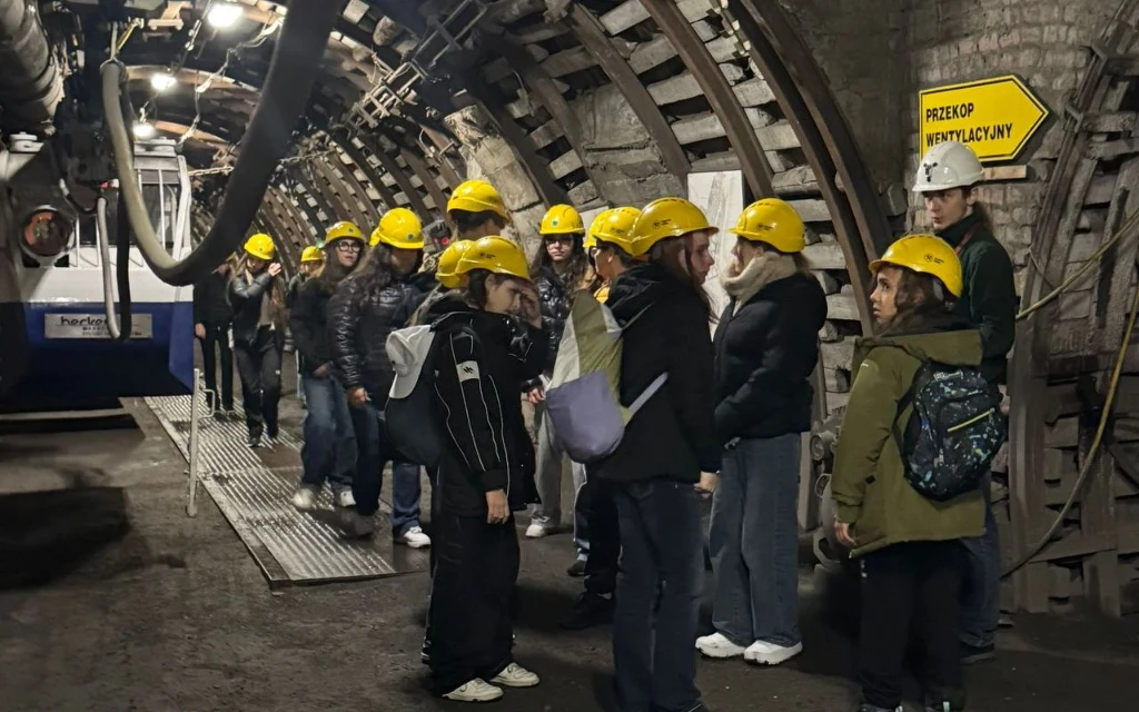Grupa osób wyposażonych w żółte kaski ochronne przechodzi przez podziemny tunel kopalni, po prawej stronie widoczny żółty znak kierunkowy z napisem „Przekop wentylacyjny”, ściany tunelu wyłożone betonowymi łukami wzmacniającymi.