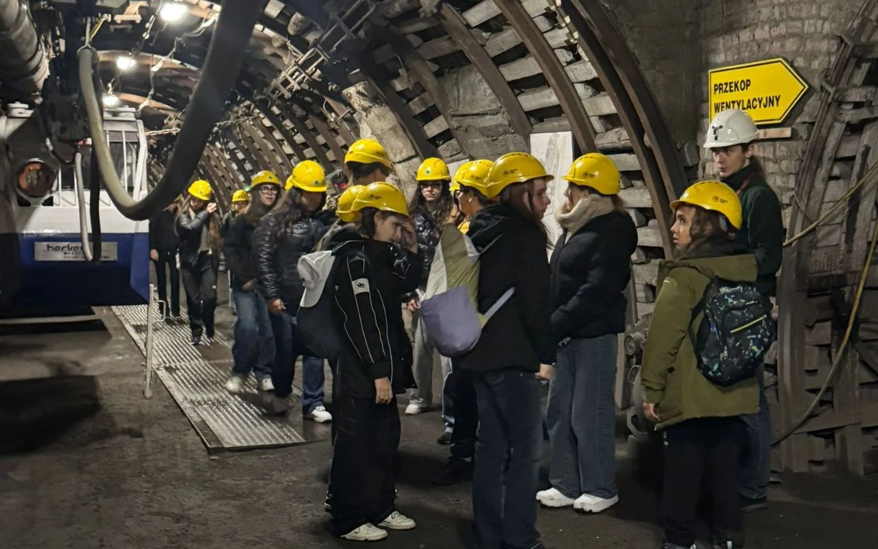 Grupa osób wyposażonych w żółte kaski ochronne przechodzi przez podziemny tunel kopalni, po prawej stronie widoczny żółty znak kierunkowy z napisem „Przekop wentylacyjny”, ściany tunelu wyłożone betonowymi łukami wzmacniającymi.