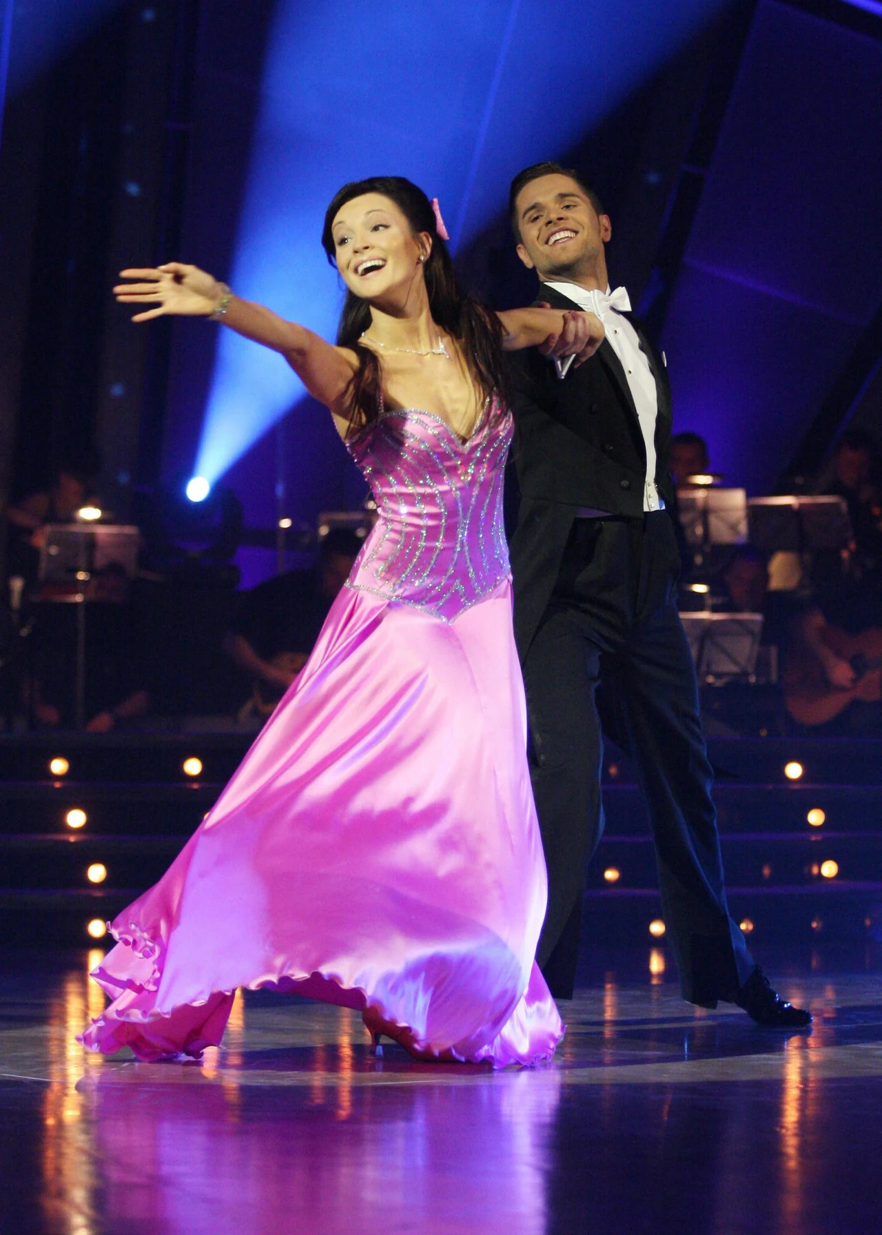 Para taneczna w eleganckich strojach wykonuje układ na oświetlonej scenie, kobieta ubrana w długą, różową suknię, mężczyzna w czarnym fraku, tło stanowi orkiestra oraz efektowne światła.