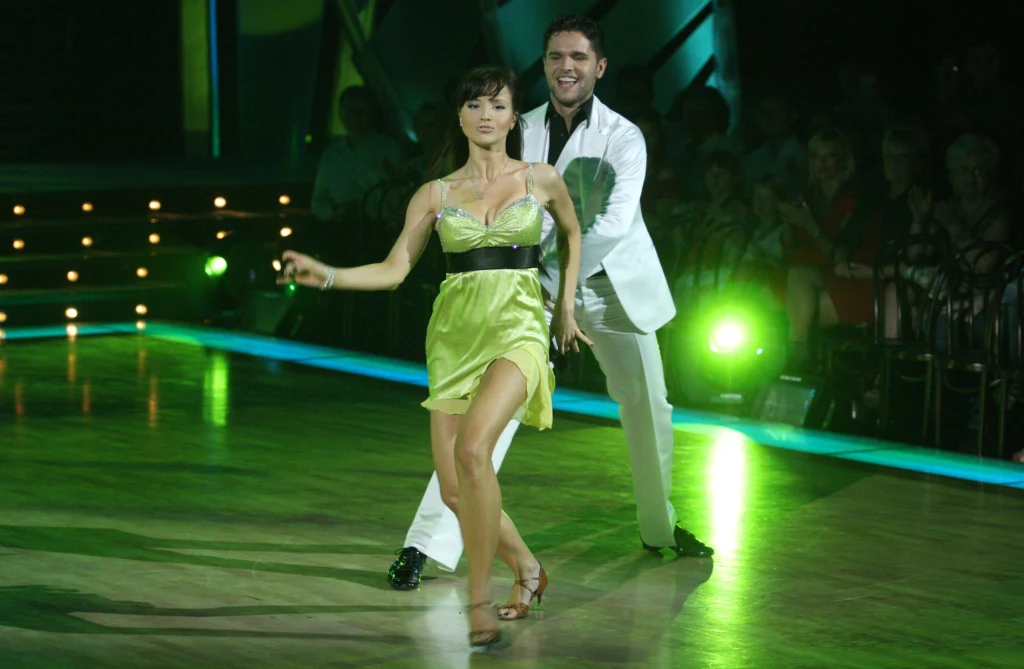 Para taneczna w eleganckich strojach tańczy na oświetlonej scenie, kobieta w jasnozielonej sukience i mężczyzna w białym garniturze uśmiechają się, wokół widoczna publiczność i światła sceniczne.