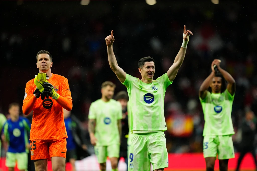 Piłkarze drużyny w jasnozielonych strojach cieszą się po meczu, jeden zawodnik unosi ręce w geście triumfu, bramkarz klaszcze, w tle widoczni są inni sportowcy i rozmyci kibice na trybunach.