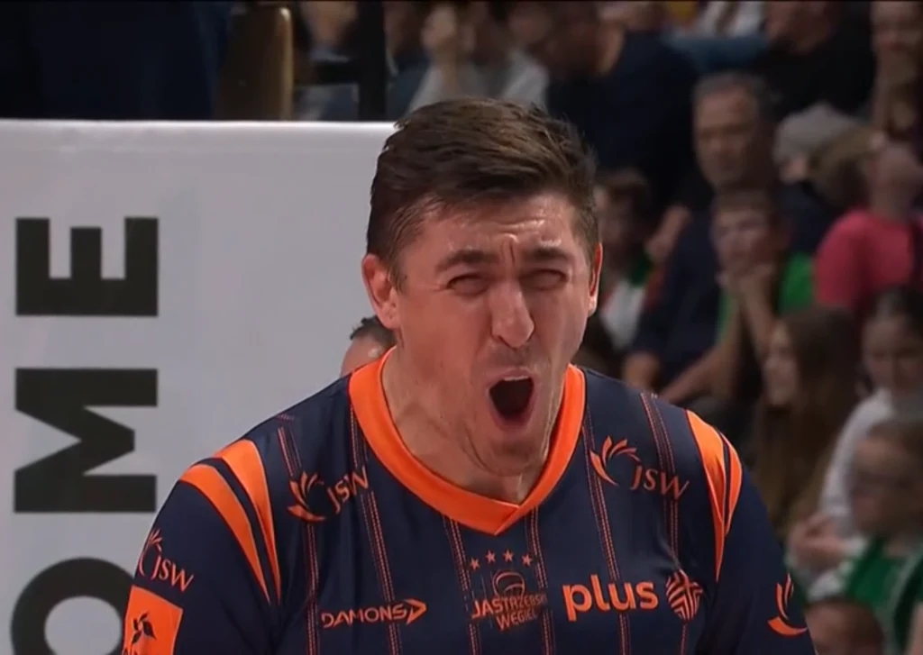 Zawodnik siatkarski w sportowym stroju z wyrazistą mimiką twarzy wyrażającą emocje, w tle rozmyta publiczność na trybunach hali sportowej.