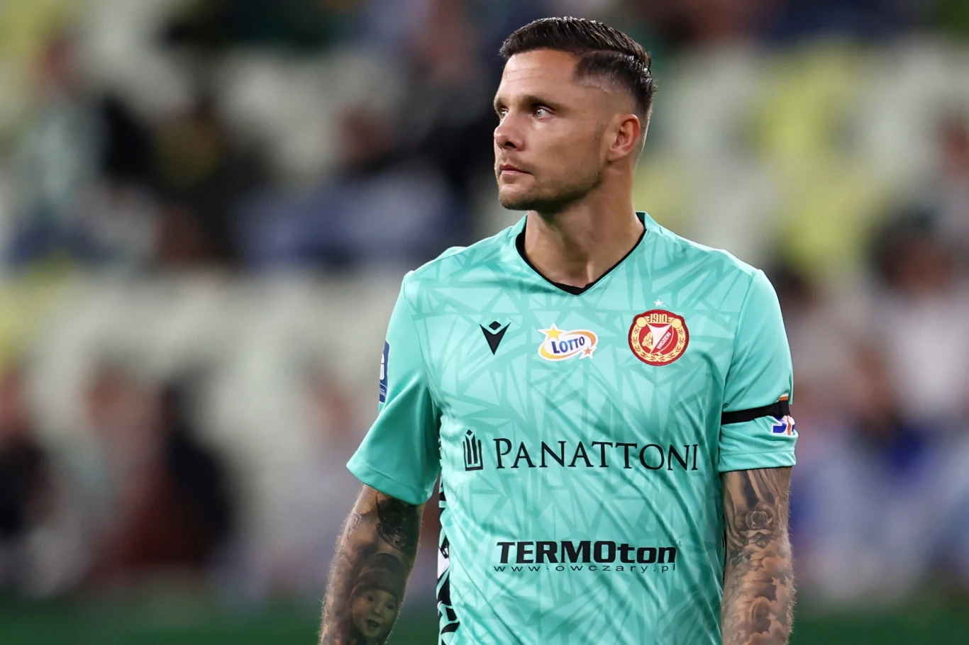 Piłkarz w zielonej koszulce z logotypami sponsorów patrzy w bok, na tle rozmytego stadionu.