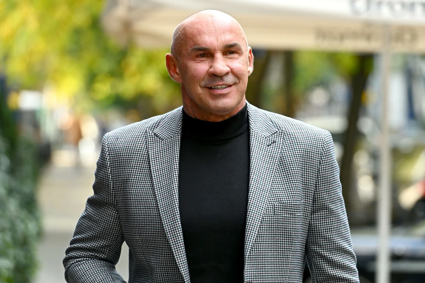 Mężczyzna w jasnej, wzorzystej marynarce i czarnym golfie stoi na tle rozmytej, zielonej przestrzeni miejskiej, uśmiechając się lekko.