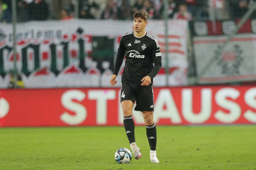 Luka Vušković Piłkarz w czarnym stroju z numerem 4 stoi na murawie podczas meczu piłki nożnej, trzymając piłkę przy nodze, w tle widoczne trybuny ze wsparciem kibiców i banerami.