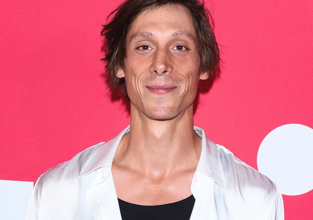 Uśmiechnięty mężczyzna o szczupłej sylwetce, ubrany w białą koszulę na czarnym podkoszulku, stoi na tle różowego ekranu z białymi elementami graficznymi.