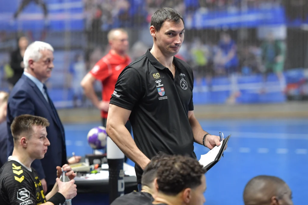 Trener drużyny piłki ręcznej w czarnej koszulce z notatkami w ręku przy linii bocznej, obok niego siedzą zawodnicy w sportowych strojach, w tle widoczni inni członkowie sztabu i elementy hali sportowej.