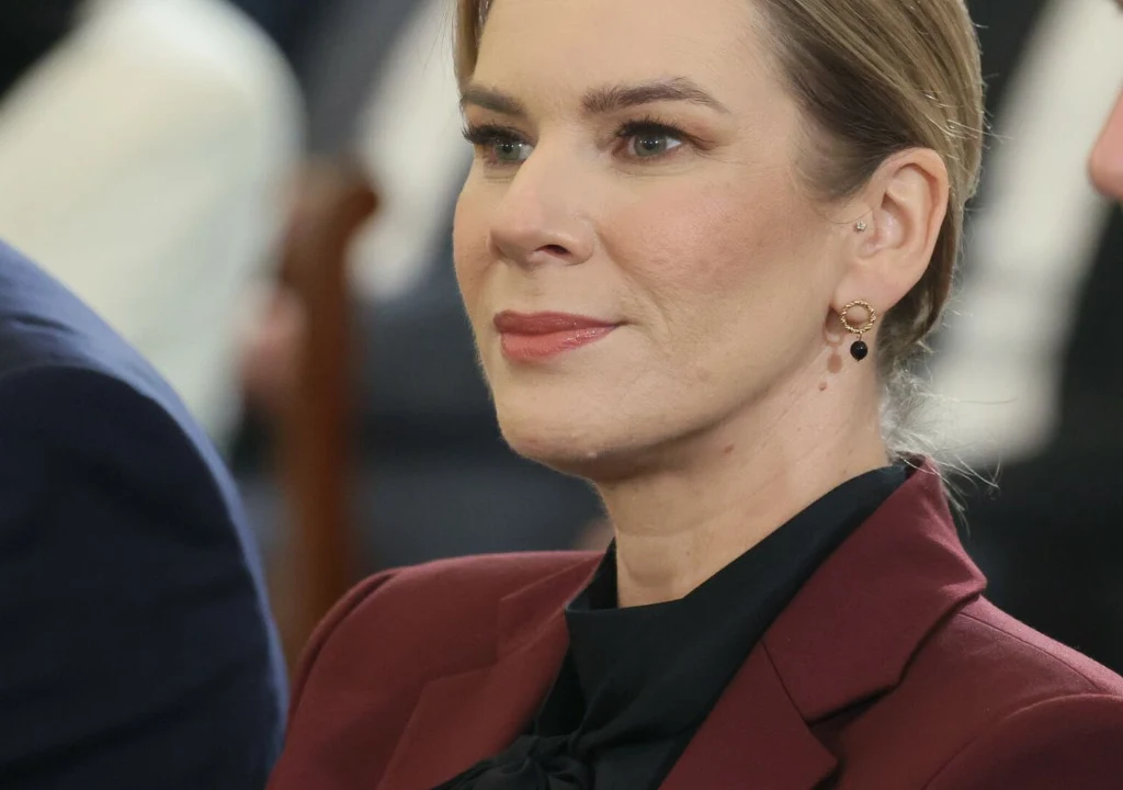 Marta Nawrocka Kobieta w bordowej marynarce i czarnej bluzce z upiętymi włosami i delikatnym makijażem, patrząca lekko w bok, w tle rozmyte sylwetki innych osób.