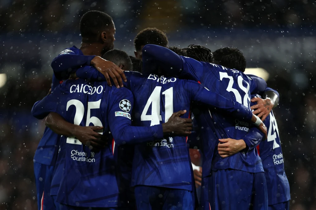 Grupa piłkarzy w niebieskich strojach obejmuje się w geście radości i wspólnoty, widoczna celebracja podczas meczu piłkarskiego w deszczu, na tle stadionu.