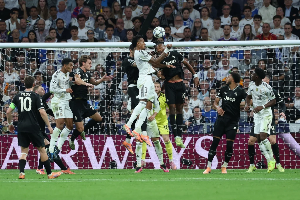 Real Madryt - Juventus (22.10.2025) Piłkarze dwóch drużyn wyskakują do piłki głową podczas dynamicznej akcji pod bramką w obecności bramkarza, na tle tłumu kibiców na trybunach.