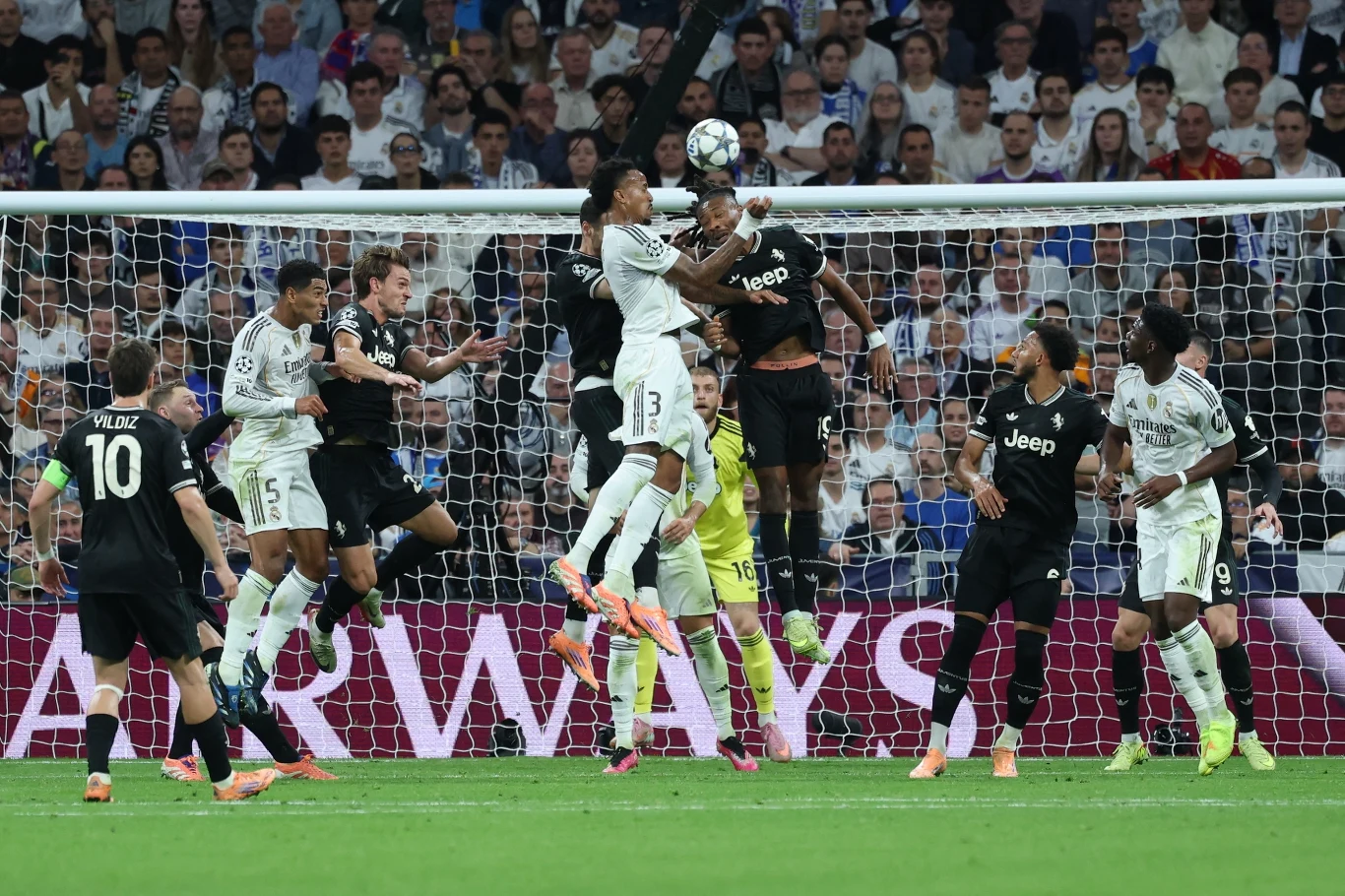 Piłkarze dwóch drużyn wyskakują do piłki głową podczas dynamicznej akcji pod bramką w obecności bramkarza, na tle tłumu kibiców na trybunach.