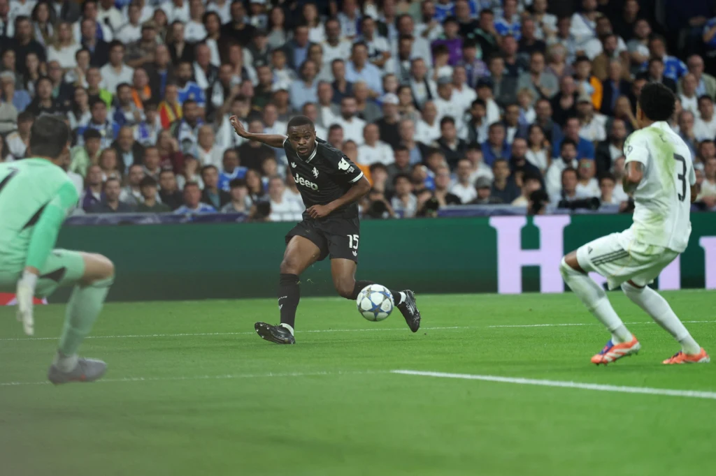 Real Madryt - Juventus (22.10.2025) piłkarz w czarnej koszulce biegnie z piłką w stronę bramki, zbliżając się do bramkarza w zielonej odzieży i obrońcy w białym stroju, na trybunach tłum widzów uważnie obserwuje akcję