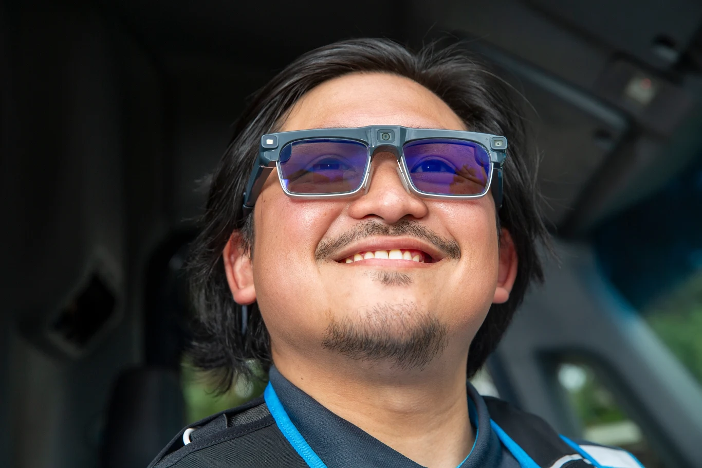 Nowe Delivery Glasses od Amazona mają polepszyć pracę kurierów Uśmiechnięty mężczyzna w okularach z niebieskimi szkłami siedzi w pojeździe, ubrany w sportową koszulkę, o ciemnych włosach i lekkim zaroście, tle rozmyte wnętrze samochodu.