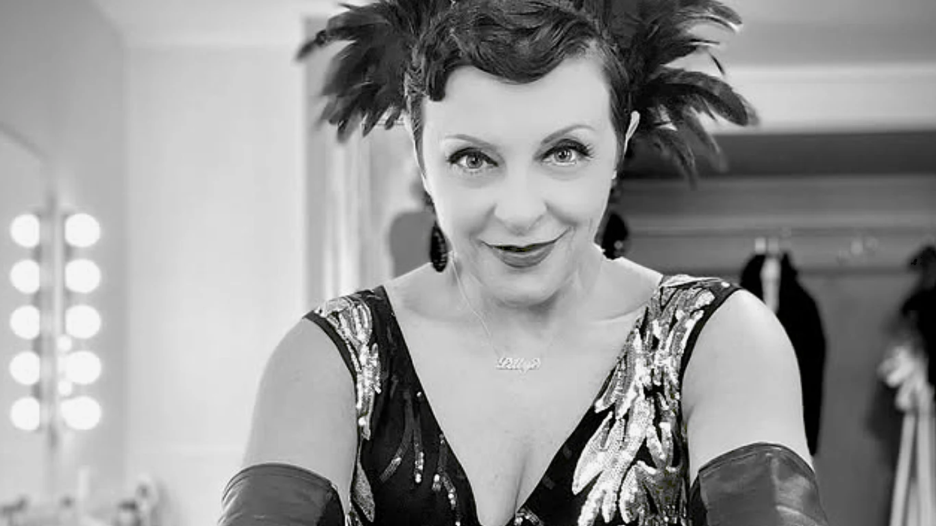 Virginia Lillye Kobieta w stylizacji retro z krótkimi, ciemnymi włosami ozdobionymi piórami, ubrana w połyskującą suknię z wzorami, w rękawiczkach, stoi przed lustrem w garderobie.