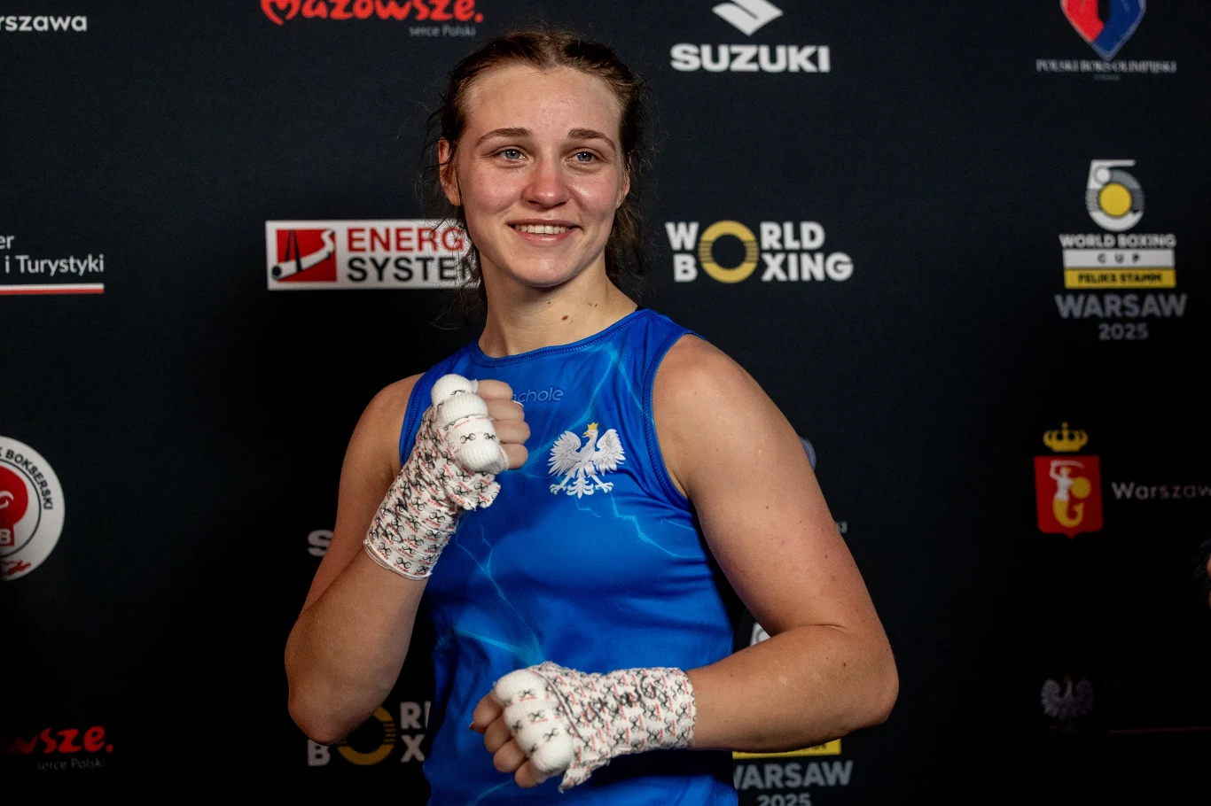 Uśmiechnięta sportsmenka w niebieskim stroju bokserskim z białym orłem na piersi, stojąca na tle ściany z logotypami sponsorów i wydarzeń bokserskich, z zaciśniętymi pięściami owiniętymi bandażami.